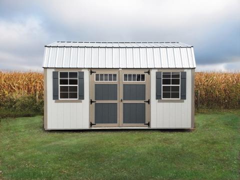 Compass High Barn shed with white siding, tan trim, white metal roofing, blue double doors, and 2 windows with blue shutters sitting in a backyard in front of a cornfield.